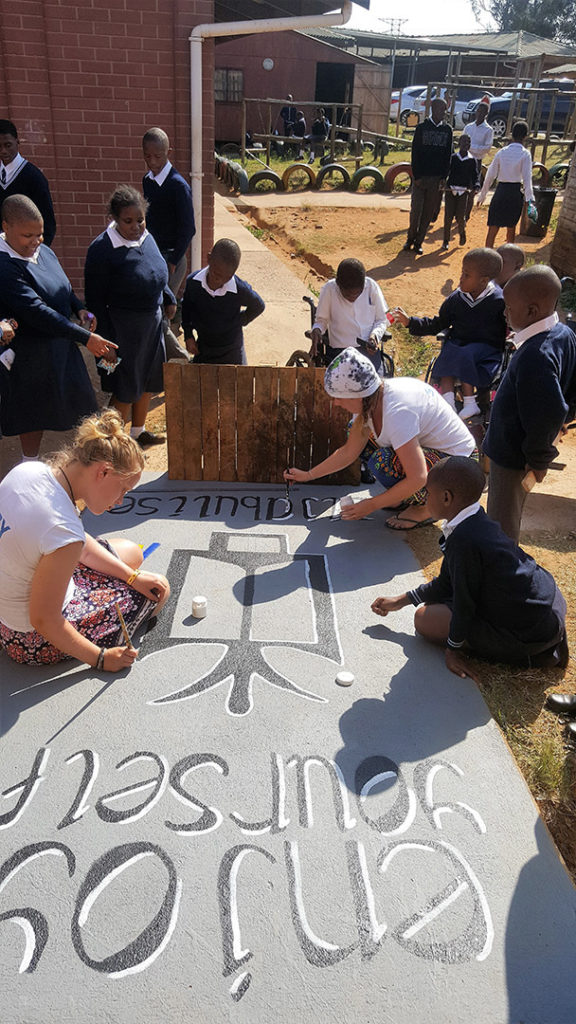 15. Mason Lincoln Art Room Entrance (b): Surrounded by learners, Lucy sits on the ramp while Suzi kneels to paint an African symbol – enjoy yourself - on the ramp.