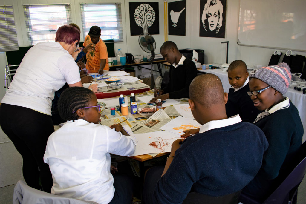 25. Mason Lincoln Braille Unit Art Class (c): Jo helps eight visually impaired learners as they sit round a table covered in supplies making tactile pictures.