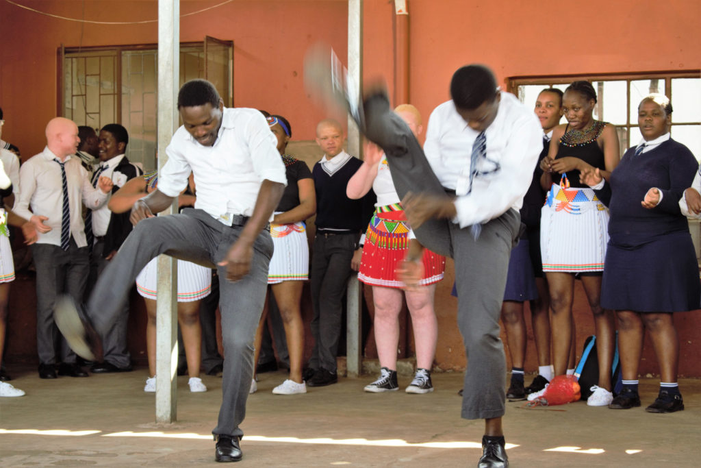 30. Mason Lincoln outside celebrating choir award (a): Two Young men in school uniform kick one leg above their heads in typical Zulu dance step to celebrate the choir’s competition as the rest of the choir look on from behind them.