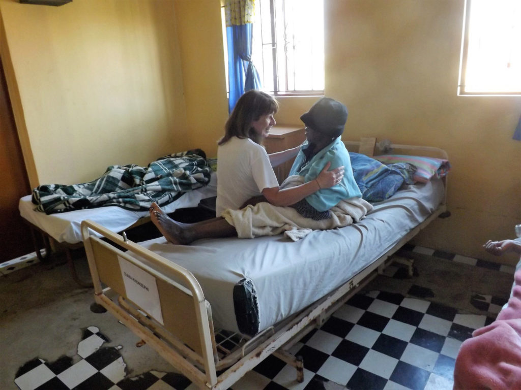 58. Ikwhezi Elderly people unit: Jean sits on hospital bed facing elderly lady with hat, they are talking and holding each other.