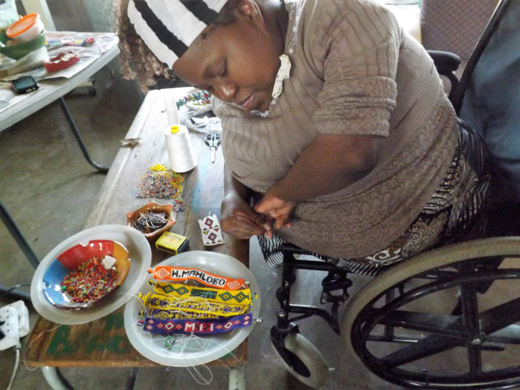 59. Ikwhezi Miriam making Zulu beads: A woman in a wheelchair bends over a table which has plates of small colourful beads and finished beadwork bracelets on it. She holds a needle and is concentrating on her work.