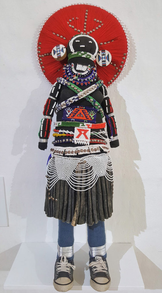 60. Art Large Makoti Doll by Lobolile Ximba self portrait of married woman passing on her wisdom about AIDS: A doll dressed in a traditional black and white costume with the addition of jeans and sneakers. She also wears a large red circular head dress.