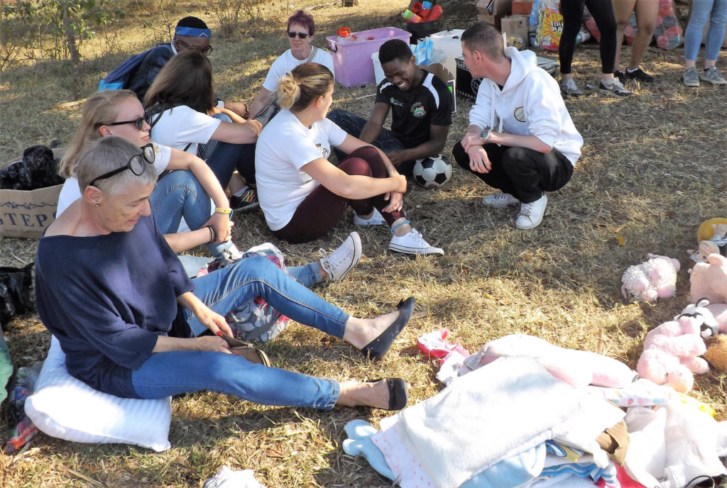 63. Bobbi Bear Under the Tree of Empowerment: The team sit on the grass under the tree, talking to one of the kids, Jackie and a social worker they are surrounded by medical supplies, clothes and toys.